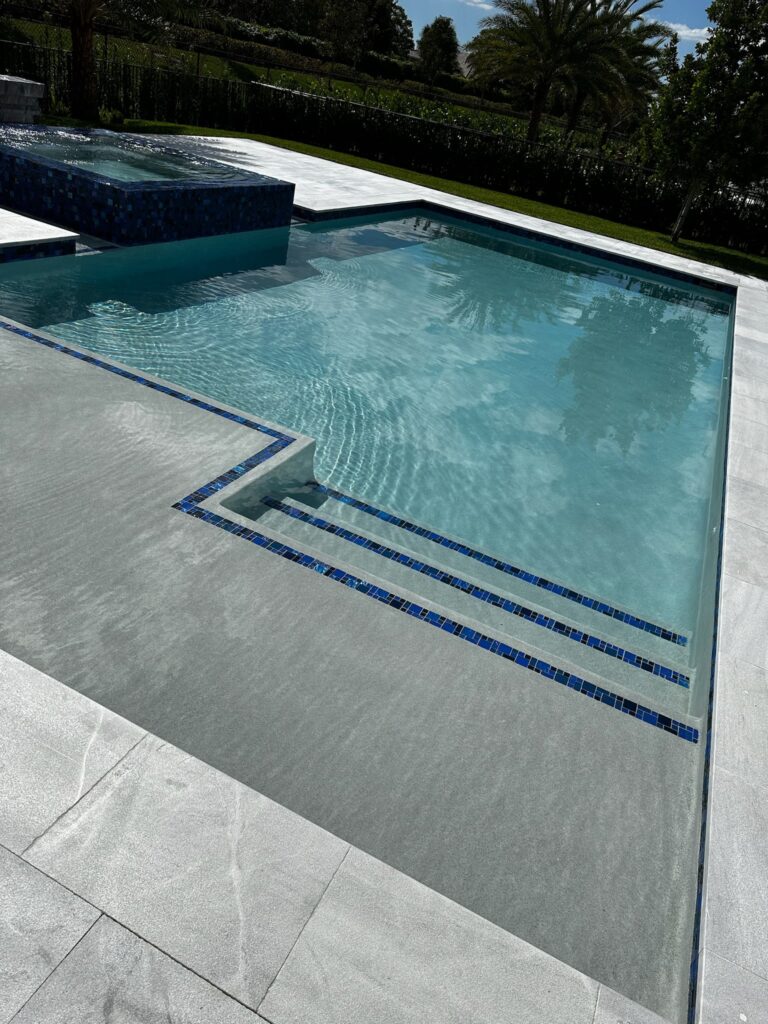 A rectangular pool with blue mosaic-tiled entry steps and a raised spa spillover, set on a travertine deck.