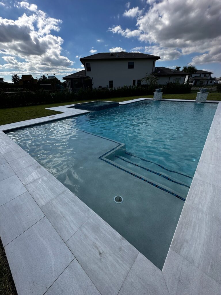 A rectangular backyard pool with a raised spa and two bubbler fountains, framed by light stone decking under a partly cloudy sky.