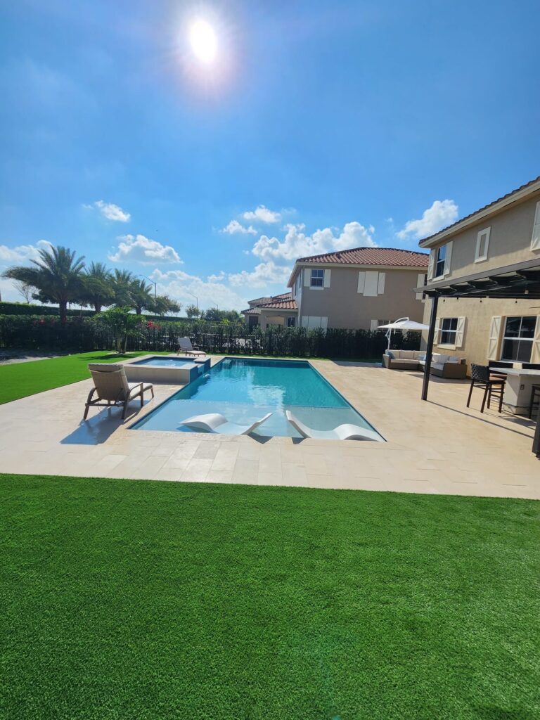 Rectangular backyard pool with raised spa, in-pool loungers, and travertine deck framed by artificial turf.