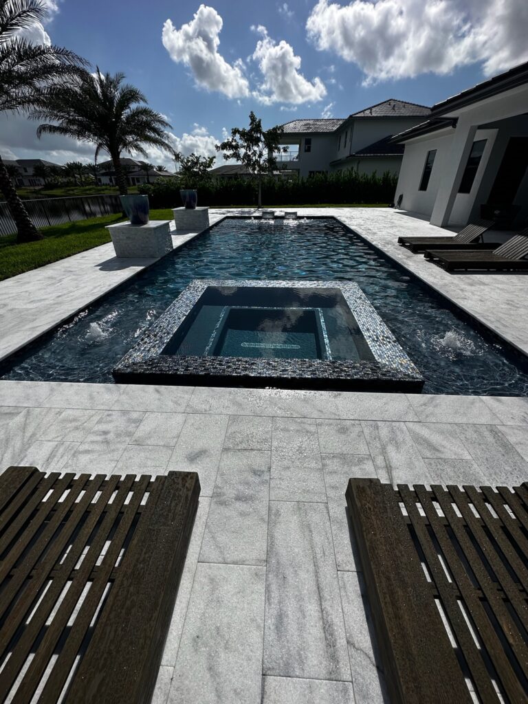 A sleek rectangular pool with an integrated glass-tile spa and bubbler jets, overlooking a canal behind a modern home.