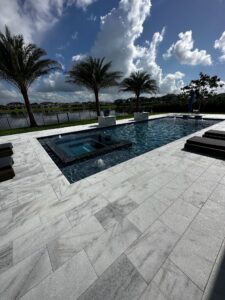 A rectangular backyard pool with a raised dark-tiled spa and water bowl fountains, set on light stone pavers with a lake and palm trees in the background.