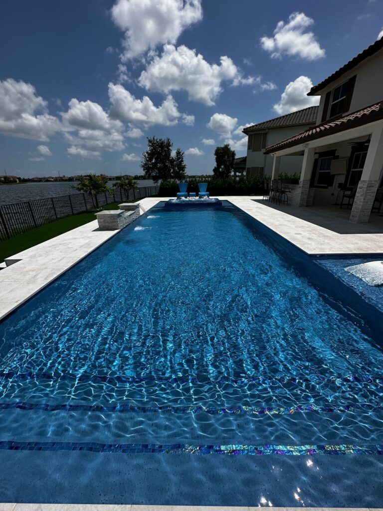 Long rectangular lakefront pool with glass tile waterline and two decorative water bowls beside a large home.