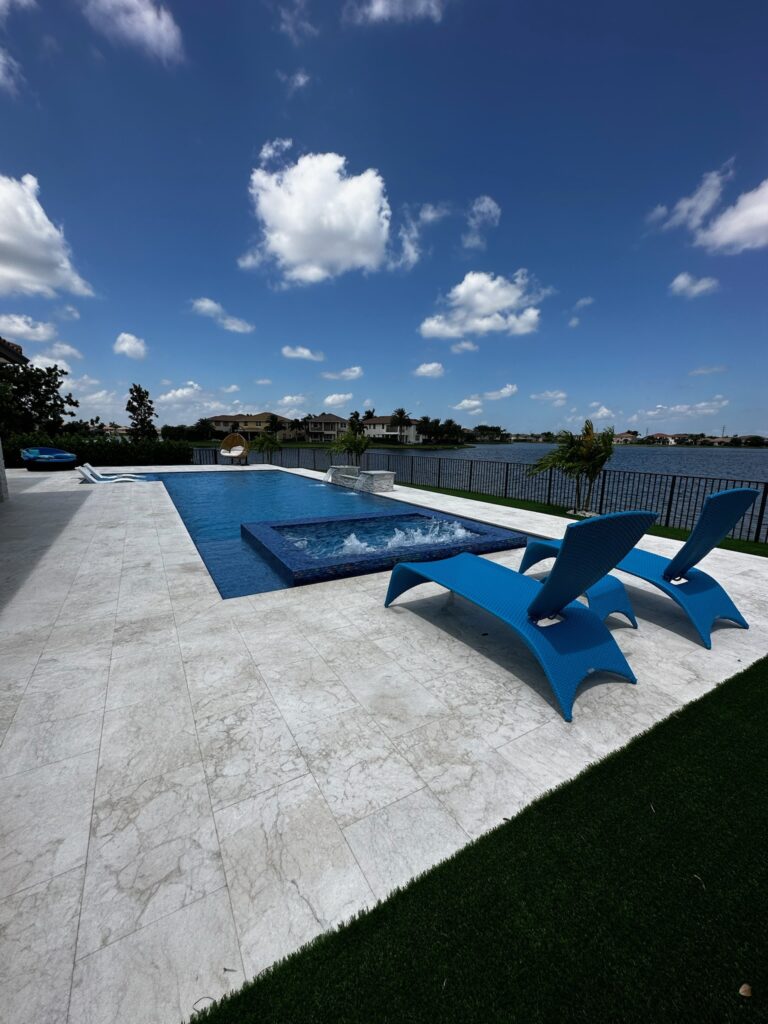 Rectangular pool with a raised spillover spa overlooking a lake, surrounded by travertine pavers.