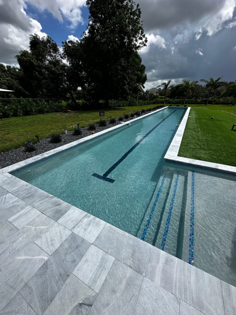 A long lap pool with blue tile lane markers and a stone deck under a stormy sky.