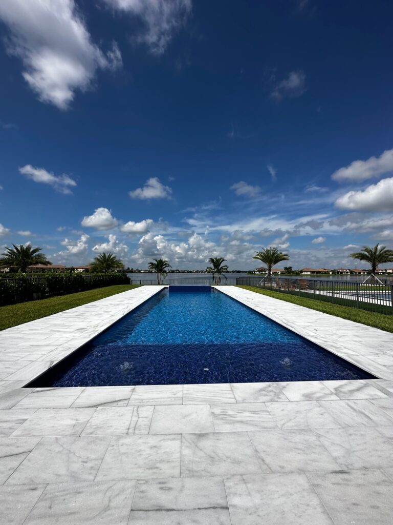 A long rectangular lap pool with a stone deck overlooking a lake under a bright blue sky.