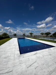 A rectangular infinity-edge style pool with dark blue tile overlooking a lake, surrounded by marble pavers.