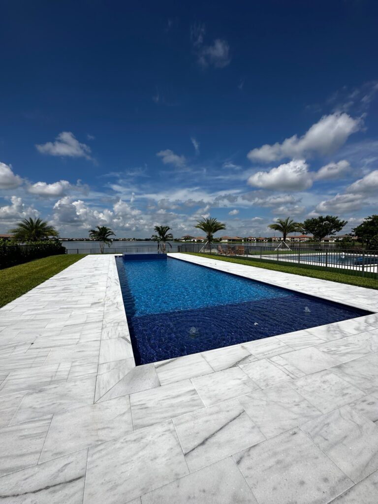 A rectangular infinity-edge style pool with dark blue tile overlooking a lake, surrounded by marble pavers.