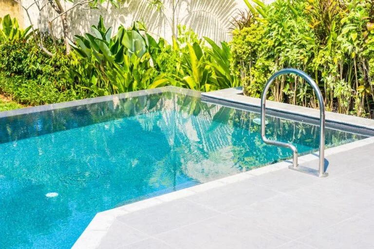 A small rectangular backyard pool with turquoise water and a stainless steel handrail, framed by tropical plants and a stone paver deck.
