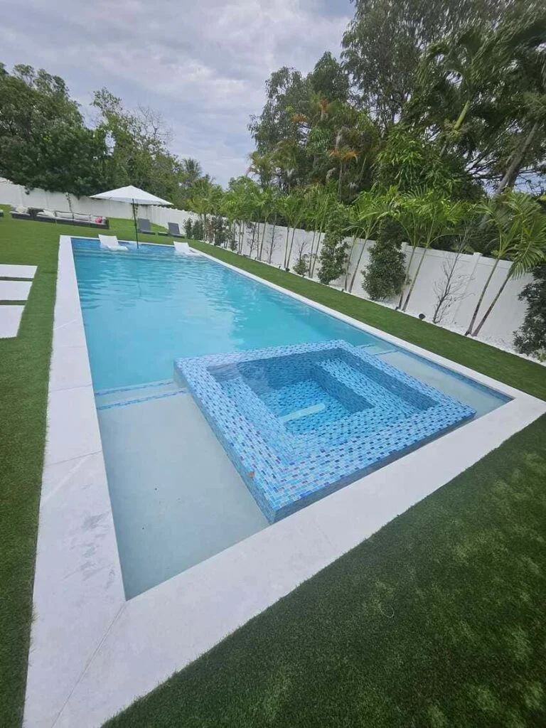 A rectangular pool with a raised blue mosaic-tiled spa at one end, surrounded by artificial turf and a white privacy wall.