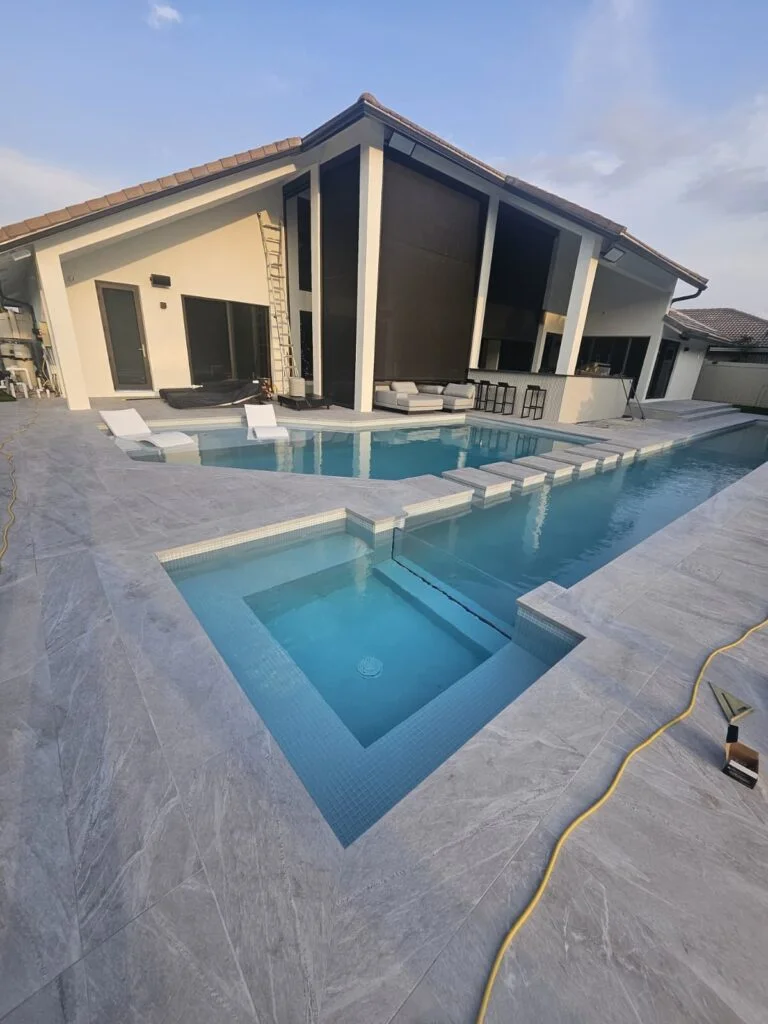 An L-shaped pool with a raised square spa and stepping-pad walkway beside a modern home under construction.