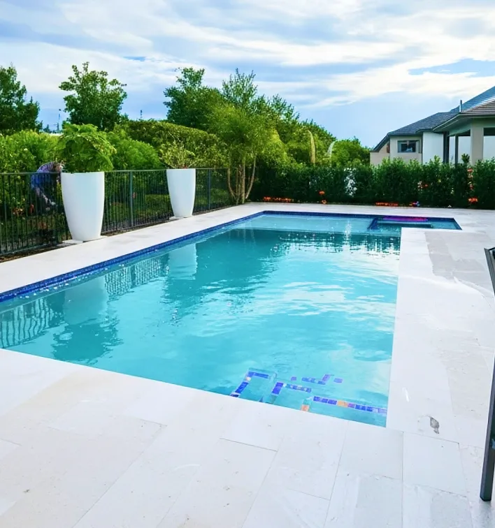 A rectangular backyard pool with a raised spa at the far end, travertine deck, and a colorful tile mosaic accent at the entry steps.