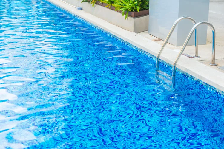 Close-up of a tiled pool edge with a stainless steel ladder and blue water.
