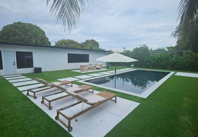 A small rectangular pool with dark water surrounded by artificial turf, pavers, loungers, and an umbrella in a suburban backyard.