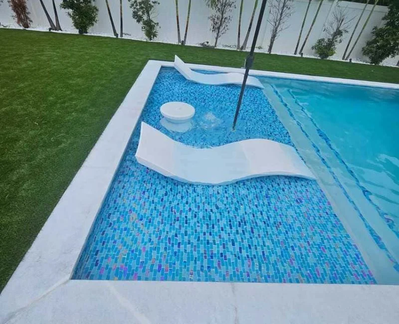 A tiled tanning ledge with two white in-pool loungers and an umbrella base at the corner of a mosaic-tiled pool.