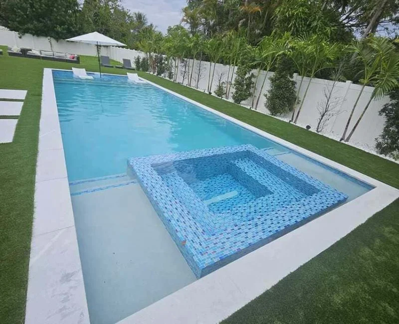 Rectangular pool with a raised blue mosaic tile spa set into the center of the far end, surrounded by artificial turf and a white privacy wall.