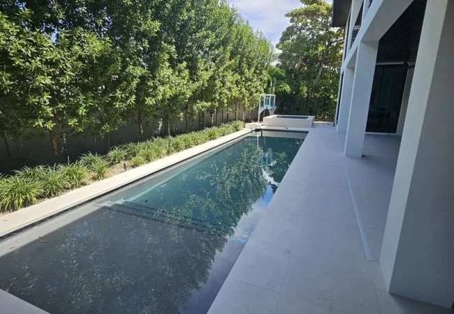 A long narrow lap pool with dark plaster runs alongside a modern white house, with a raised spa at the far end.