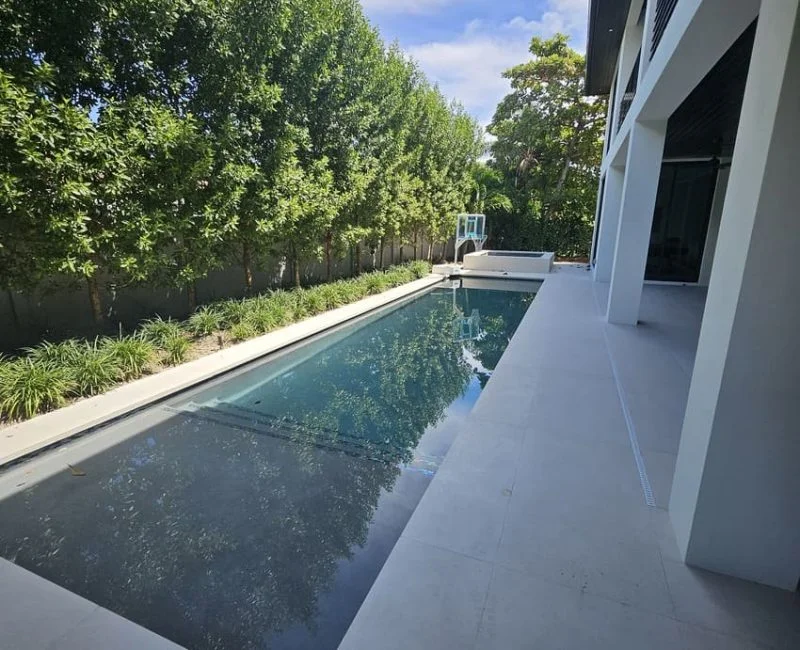 A long narrow lap pool with dark water runs alongside a modern home, with a raised spa at the far end.