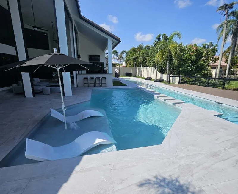 A long geometric pool with in-water loungers on a tanning ledge, framed by a modern house and palm trees.