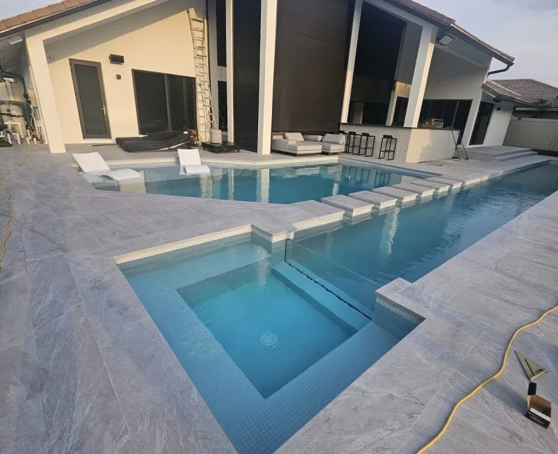 A modern multilevel pool with a raised spa and stone paver deck beside a house at dusk, with construction materials visible nearby.