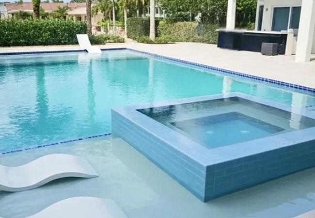 A rectangular backyard pool with a raised square tiled spa and white lounge chairs on a stone patio.