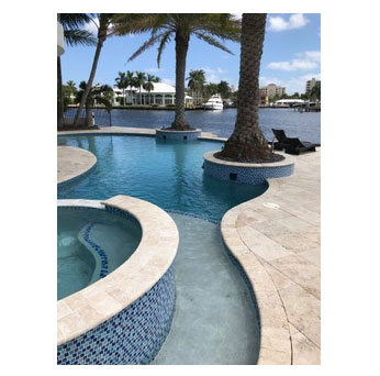 Freeform pool with raised blue-tiled spa and travertine deck overlooking a waterway lined with palm trees.
