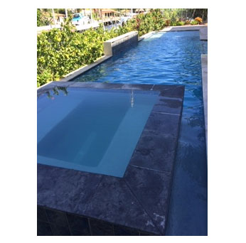 A raised square spa with a small spout sits beside a long narrow lap pool overlooking a waterway.
