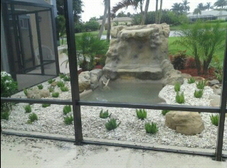 A small rock waterfall feature surrounded by white gravel landscaping, seen through a screened enclosure.