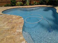 A small freeform backyard pool with travertine coping and a cleaning hose in the water.