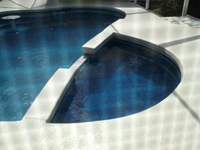 A corner view of a pool with a curved raised wall and light-colored paved deck.