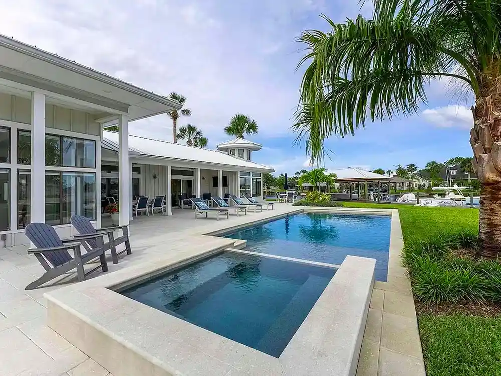 Rectangular pool with an elevated spa on a stone patio beside a waterfront home with palm trees.