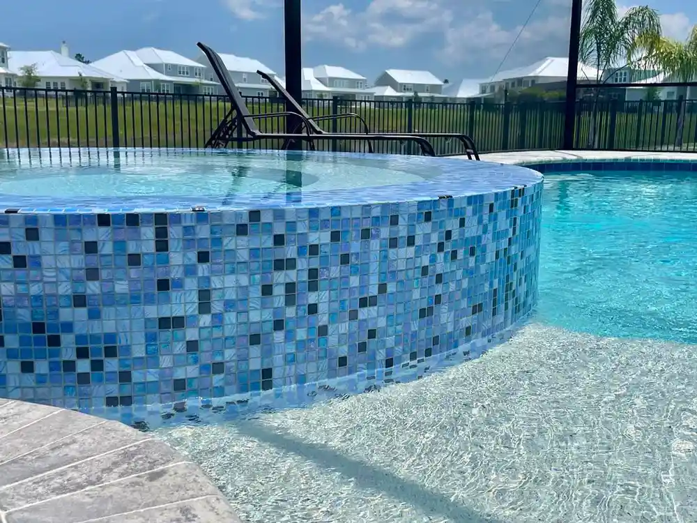 A raised round spa covered in blue mosaic tile sits beside a pool with a backyard fence and neighboring houses beyond.