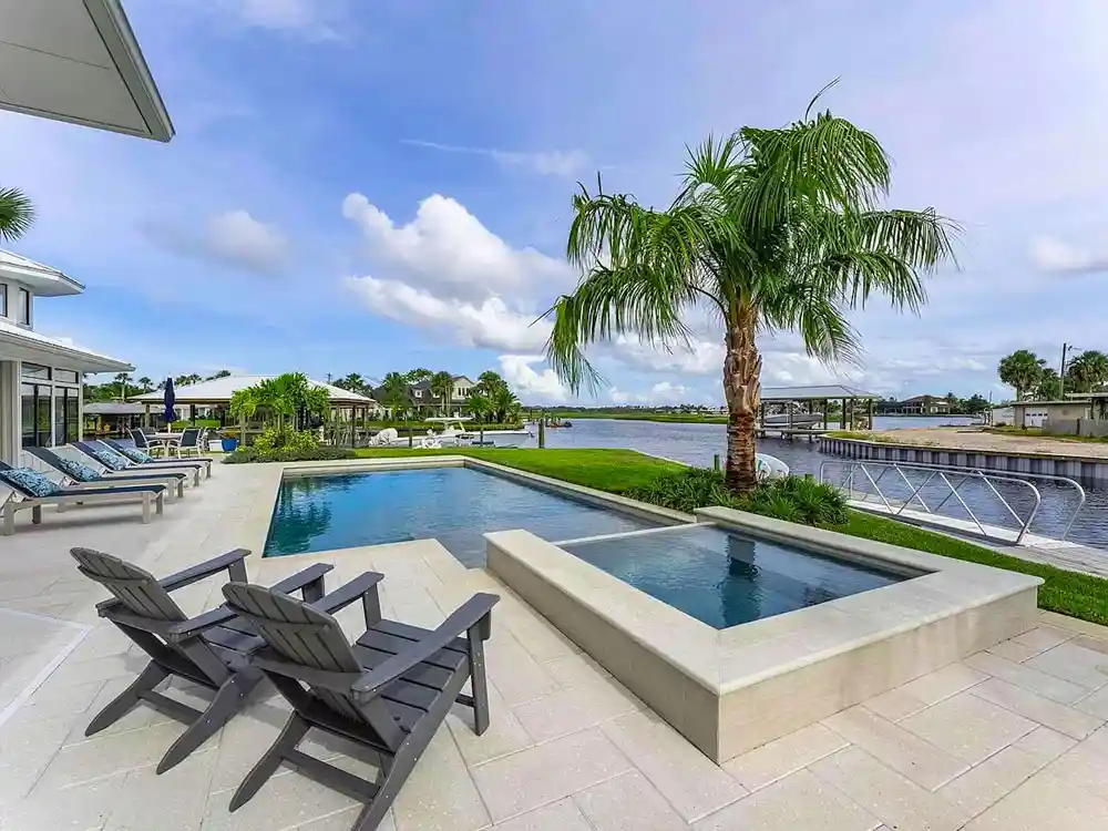 A rectangular pool with a raised spa sits beside a canal, framed by a large palm tree and lounge chairs.