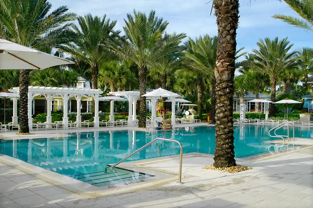 A large resort-style rectangular pool surrounded by palm trees, pergolas, and lounge chairs.
