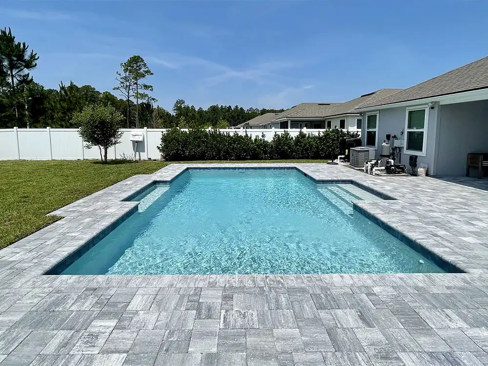 A rectangular in-ground pool with gray paver decking in a fenced suburban backyard.