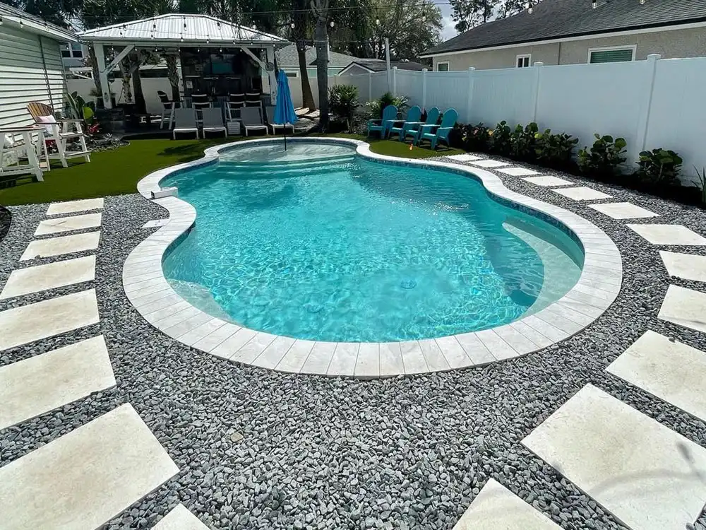 A freeform fiberglass pool with an integrated raised end and paver-and-gravel patio in a fenced suburban backyard.