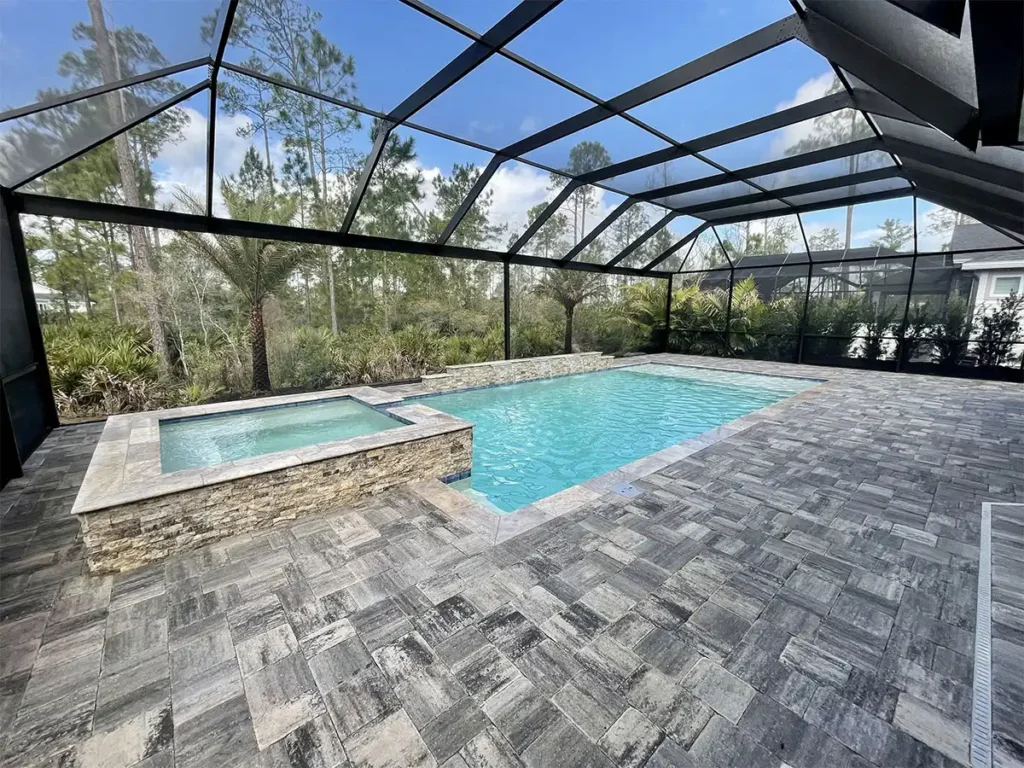 A rectangular pool with a raised stacked-stone spa under a screened lanai surrounded by trees.