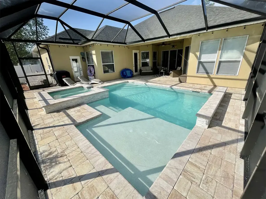 Screened backyard pool with an L-shaped tanning ledge and an attached raised spa on a travertine paver deck.