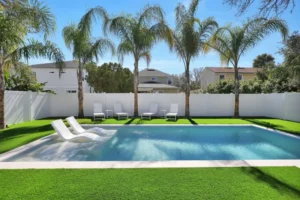 A small rectangular backyard pool with a submerged tanning ledge and white loungers, surrounded by artificial turf and palm trees.