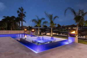 A raised glass-tiled spa glows blue at night beside a canal-front pool with lit fire bowls on stone pedestals.