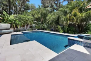 A rectangular backyard pool with a tiled raised spa edge surrounded by lush tropical palms and a travertine deck.