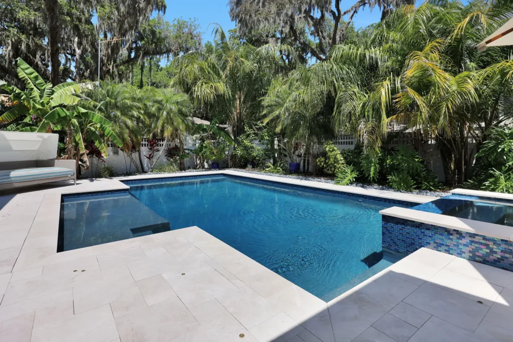 A rectangular backyard pool with a tiled raised spa edge surrounded by lush tropical palms and a travertine deck.