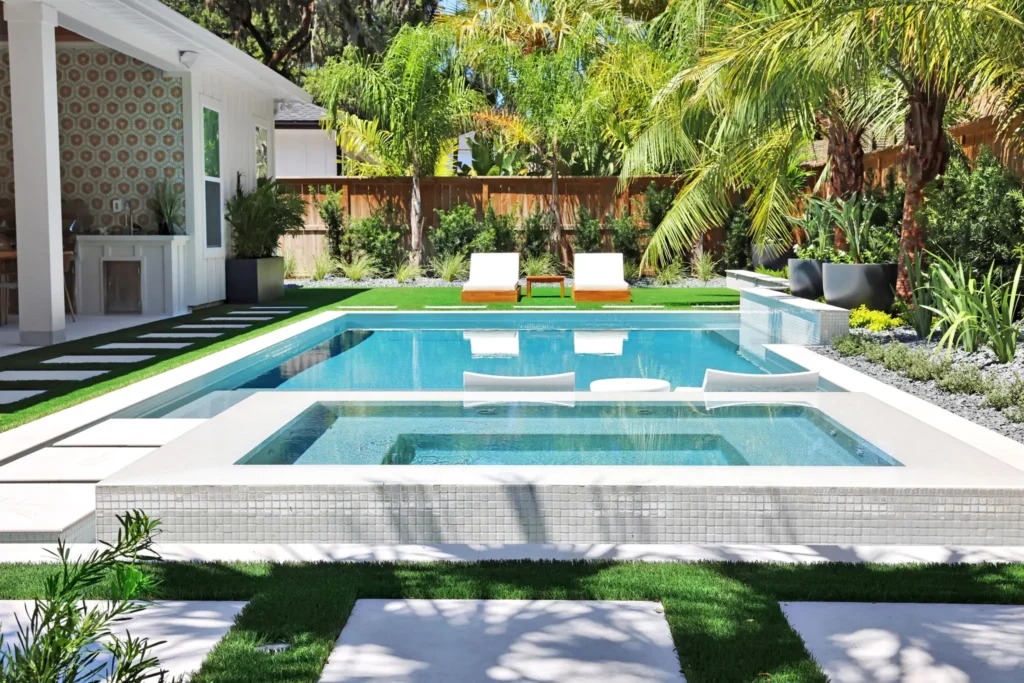 A rectangular pool with a raised tiled spa at one end, floating loungers, and a turf and paver backyard lined with palms.
