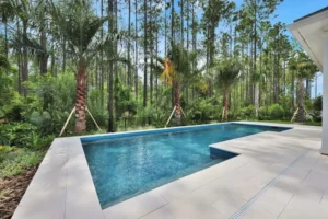 A rectangular backyard pool with blue tile waterline and stone deck set against a wooded tree line.