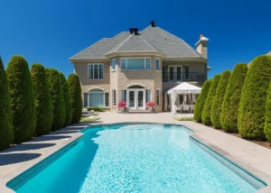 A rectangular backyard pool with turquoise water in front of a large stone mansion flanked by tall manicured hedges.