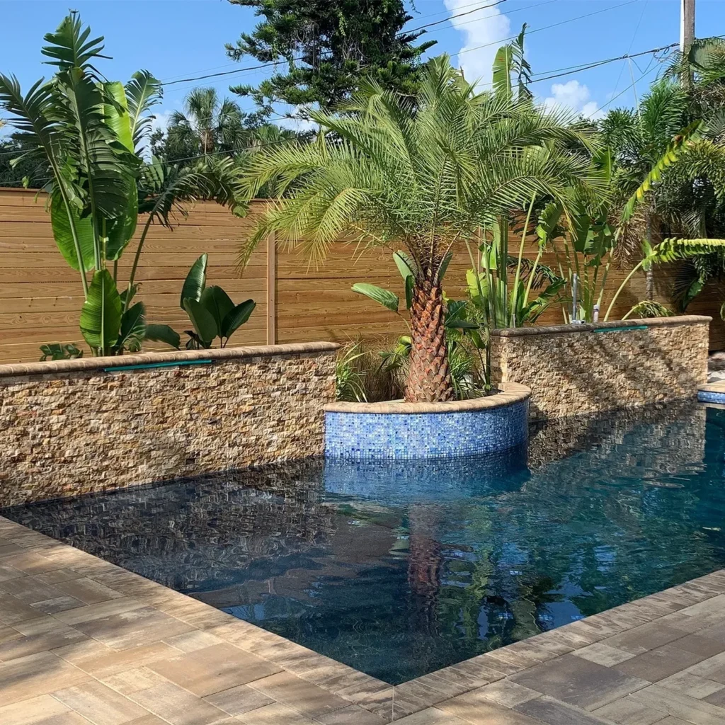A pool corner with a raised stone-clad planter topped with mosaic tile and a large palm tree, surrounded by paver decking.