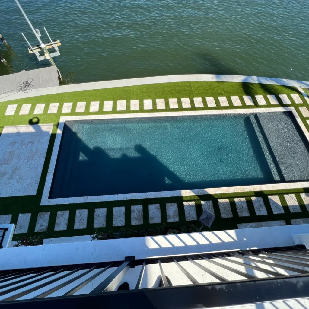 Aerial view of a rectangular pool with a dark accent section beside a canal-front lawn with turf and stone pavers.