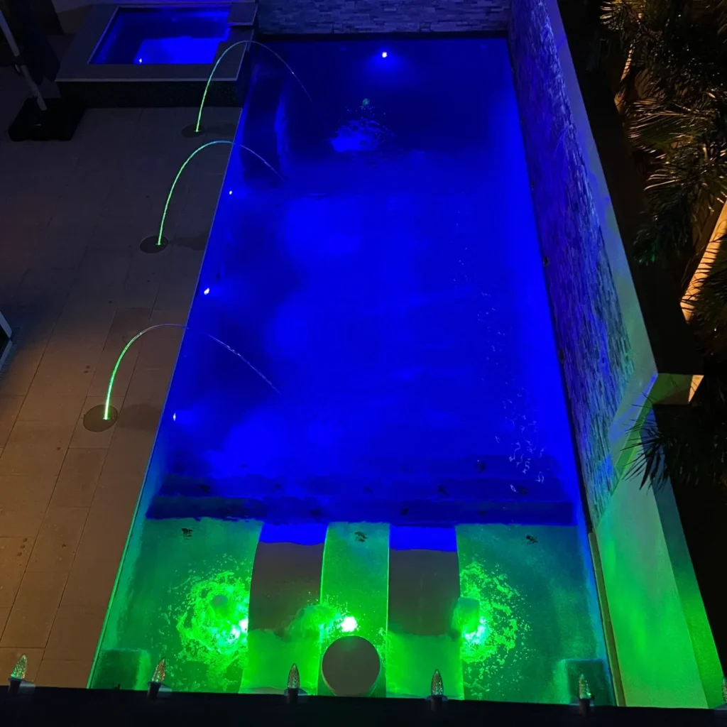 An aerial night view of a rectangular pool lit in blue and green LED light with fountain jets and a raised spa.