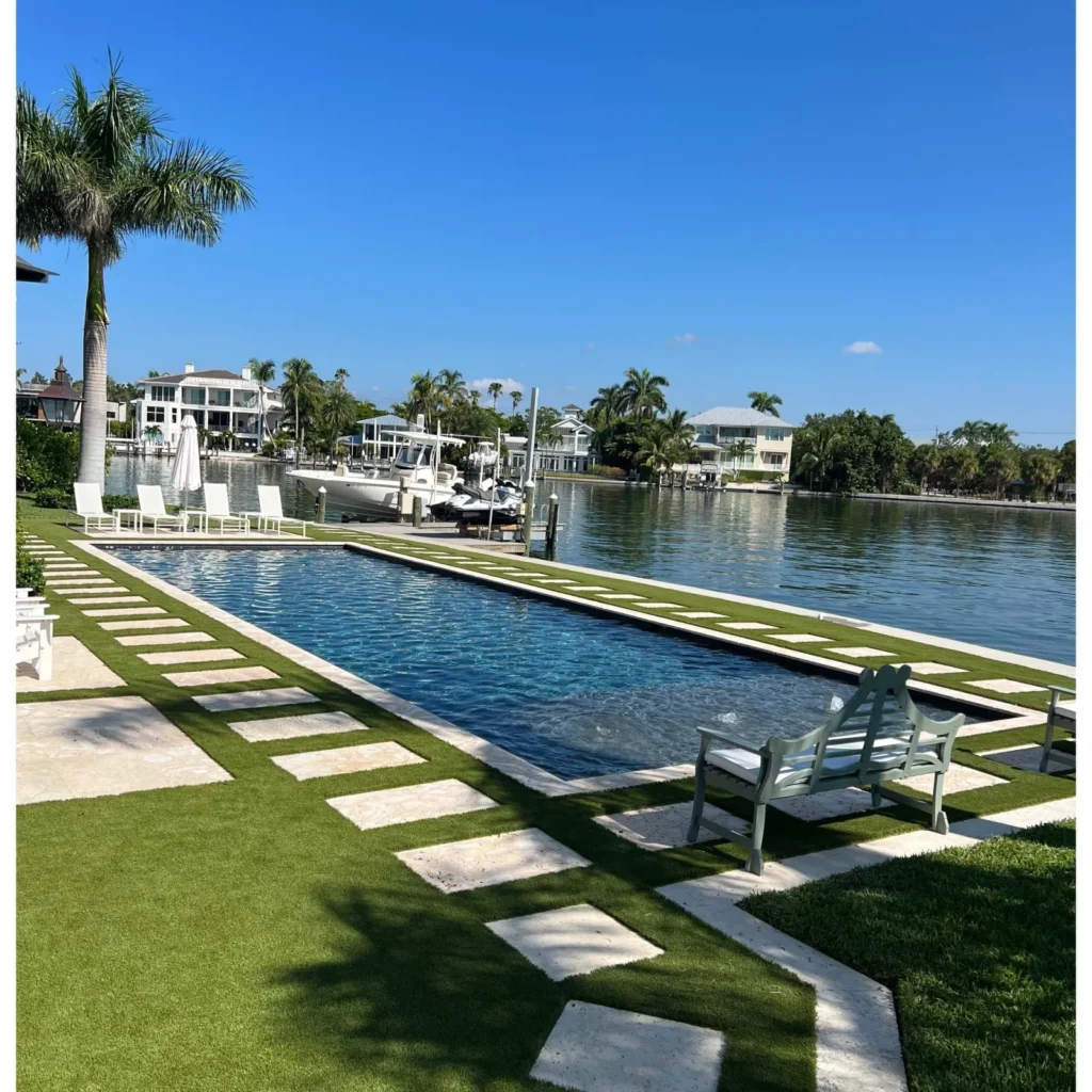 A long narrow lap pool along a canal with turf and travertine paver decking and a bench.