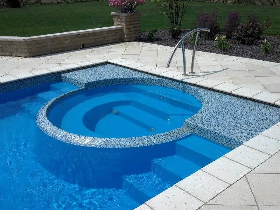 A round tiled spa with steps sits flush with a rectangular pool on a travertine patio.
