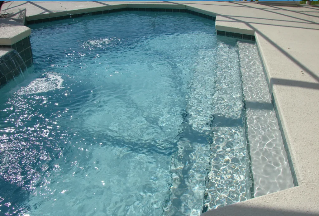 Close view of a pool corner with wide entry steps and a small tiled waterfall feature under a screened enclosure.